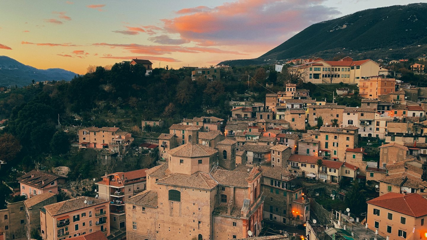Città europea con tetti in terracotta su una collina boscosa al tramonto.